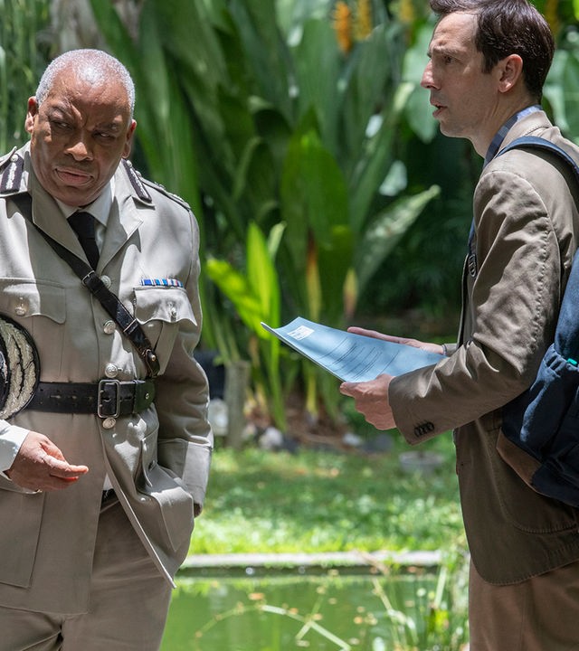 "Death in Paradise - Die letzte Botschaft (2):" Commissioner Selwyn Patterson (Don Warrington) steht im Grünen und blickt mürrisch zur Seite, während DI Neville Parker (Ralf Little) mit einer Polizeiakte in den Händen neben ihm steht und sprachlos in seine Richtung schaut.