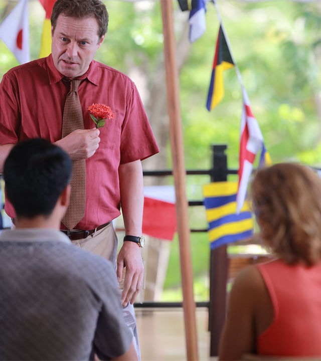 "Death in Paradise - Geldgier": DI Jack Mooney (Ardal O''Hanlon) hält eine Blume in der Hand, im Hintergrund sind verschiedene Länderflaggen zu erkennen.