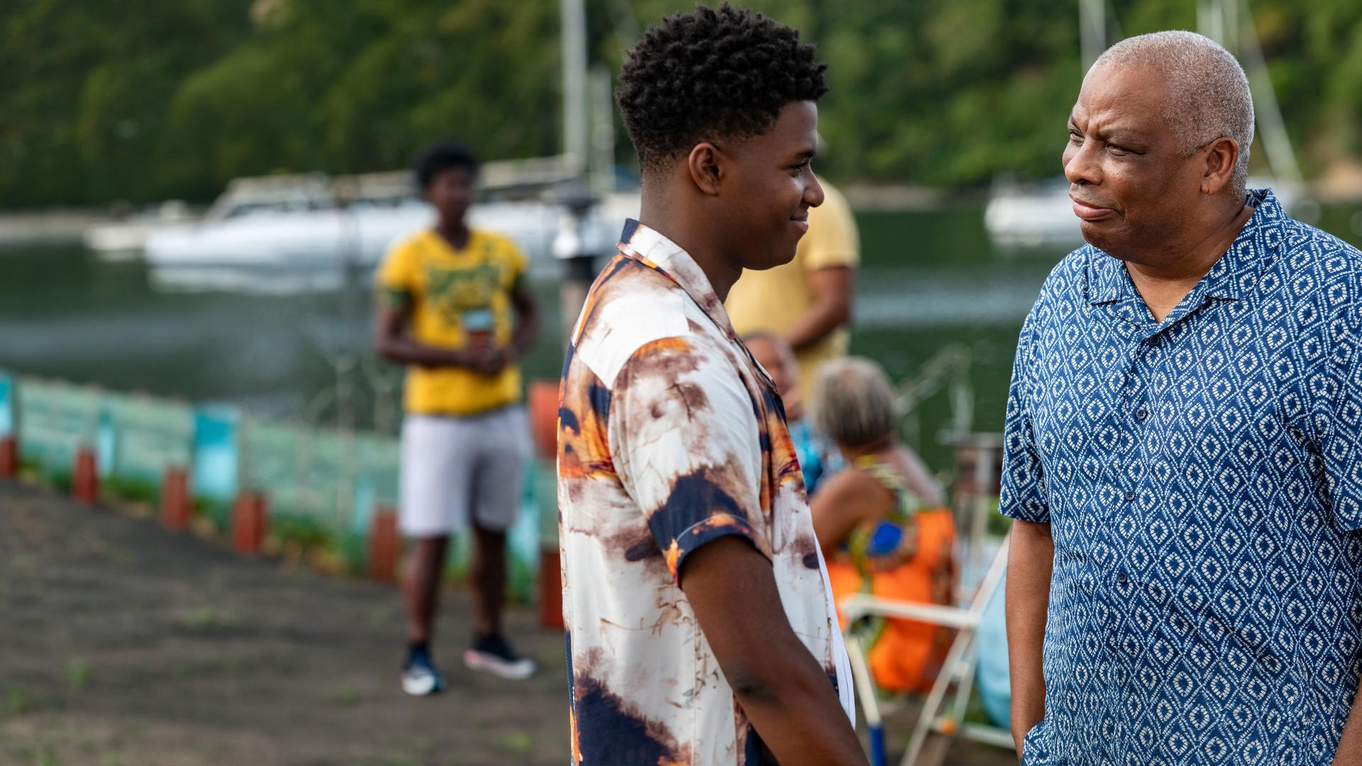 "Death in Paradise - Licht im Dunkeln": Marlon (Tahj Miles) und Commissioner Patterson (Don Warrington) stehen sich auf einem Pier gegenüber. Beide tragen bunte Freizeithemden und schauen sich aufrichtig an.