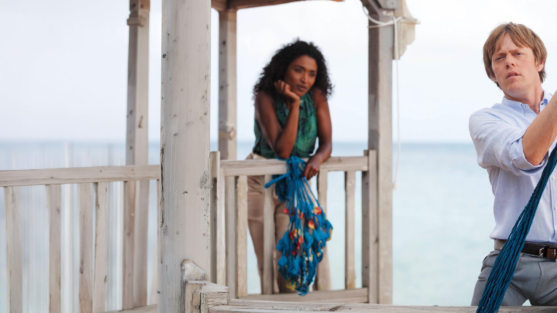"Death in Paradise - Tödliche Schokolade": Camille (Sara Martins) lehnt sich auf ein Geländer einer Hütte am Strand und schaut Humphrey an. Auf einem Arm stützt sie sich ab und mit der anderen Hand hält sie ein blaues Seil fest. Humphrey (Kris Marshall) befestigt ein Seil an einem Stamm und schaut nach vorn.