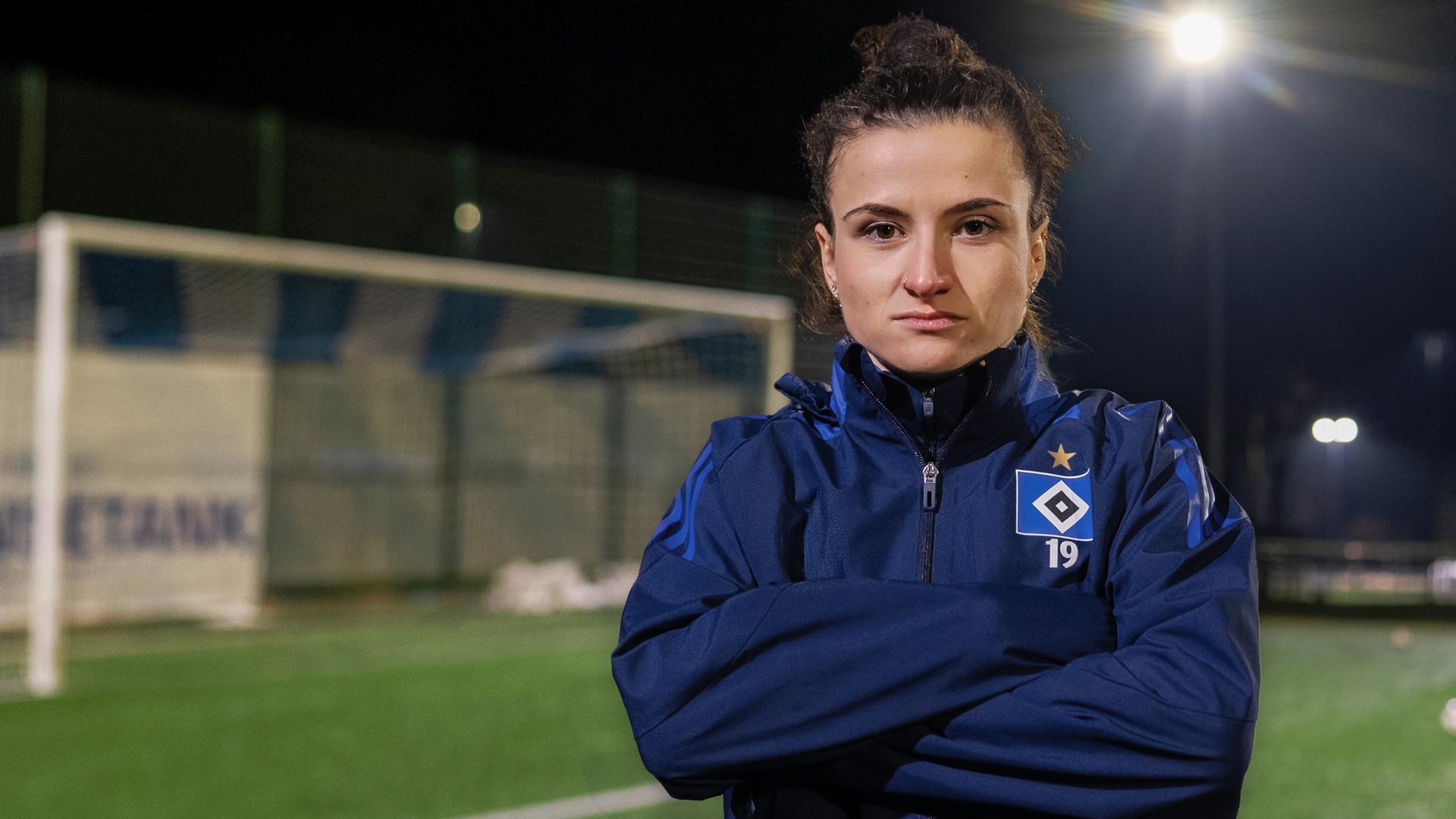 Debbie trägt einen Trainingsanzug. Sie hat die Arme vor der Brust verschränkt und schaut starr in die Kamera. Im Hintergrund ist zu erkennen, dass sie auf einem Fußballplatz steht, da links hinter ihr ein Tor zu sehen ist.  