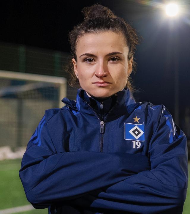 Debbie trägt einen Trainingsanzug. Sie hat die Arme vor der Brust verschränkt und schaut starr in die Kamera. Im Hintergrund ist zu erkennen, dass sie auf einem Fußballplatz steht, da links hinter ihr ein Tor zu sehen ist.  