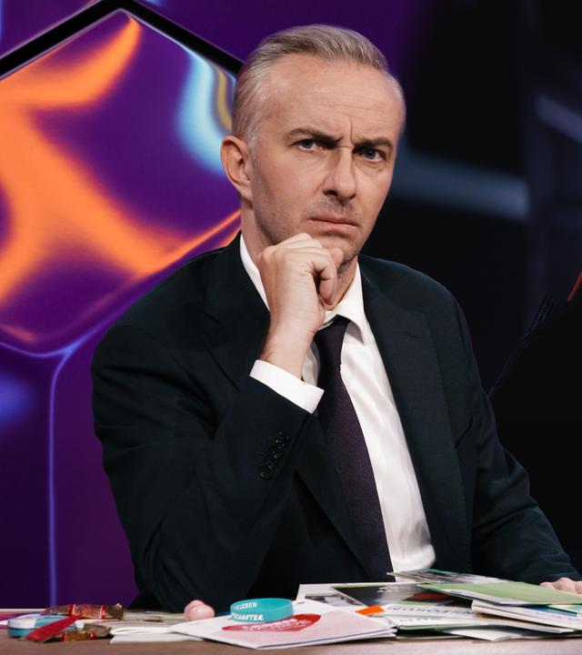 Jan Böhmermann sitzt am Schreibtisch. Rechts im Hintergrund ist eine Person mit einem Fernglas zu sehen. Auf den leuchtenden Linsen des Fernglases befindet sich das Symbol einer stilisierten Gebärmutter.