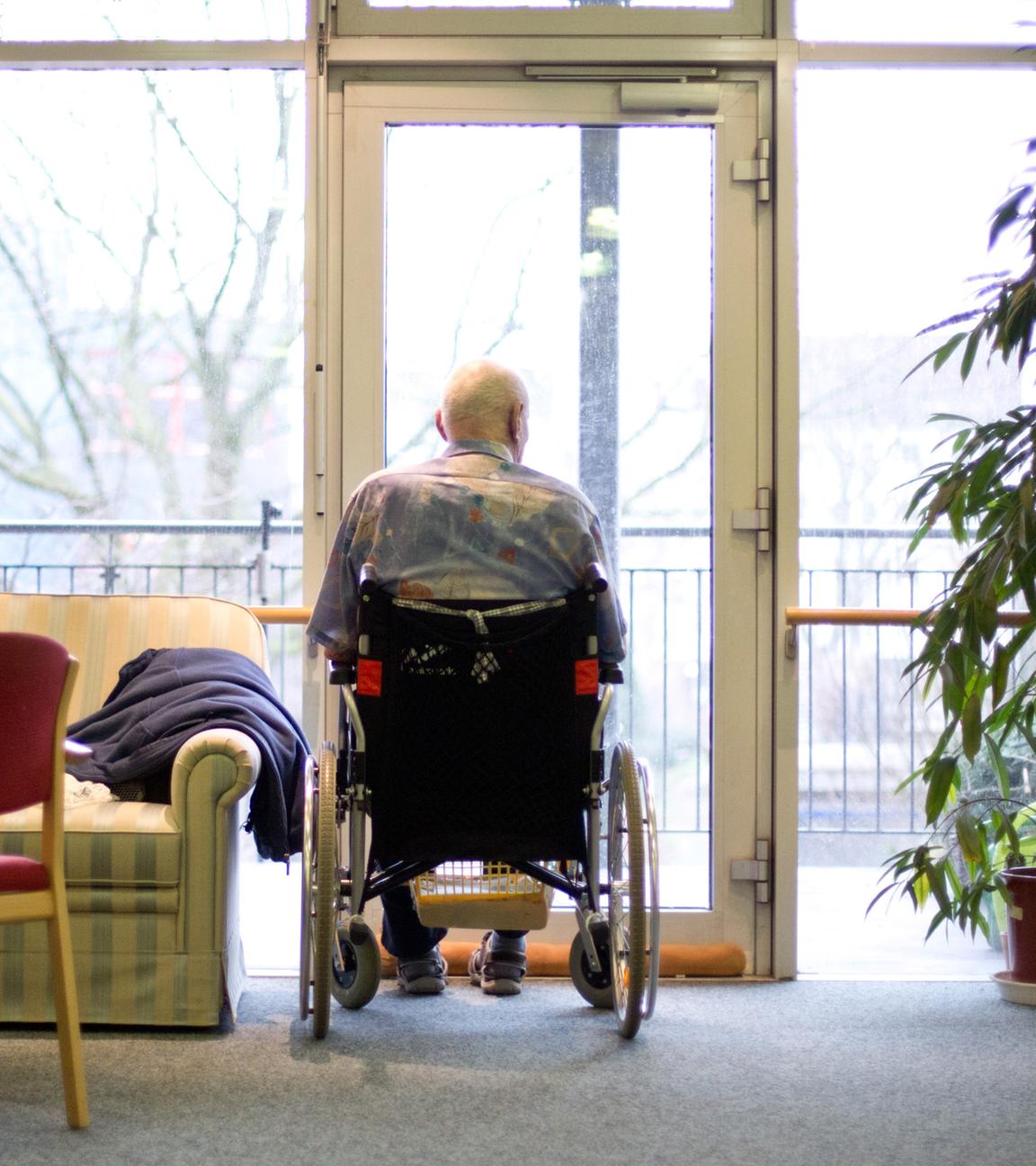 Ein alter Mann sitzt im Rollstuhl am Fenster und blickt heraus.