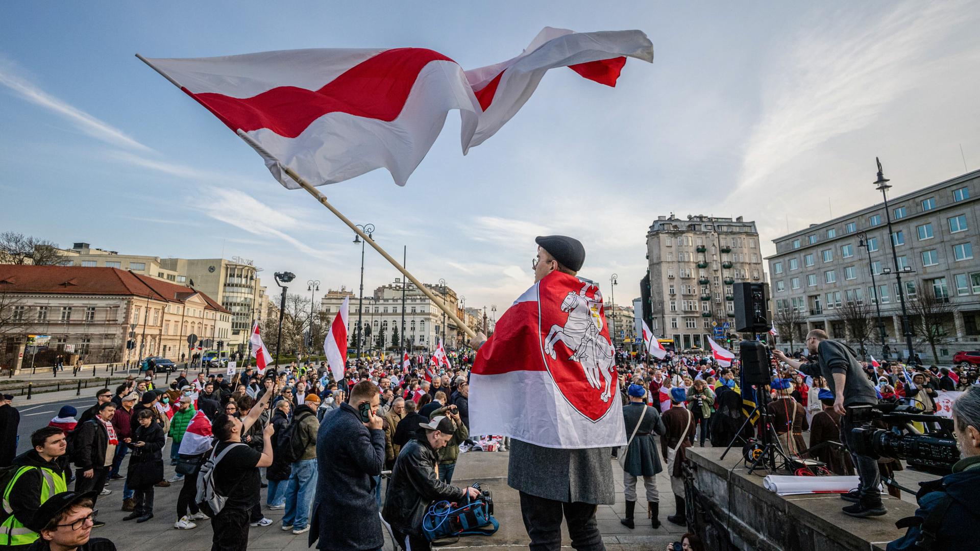 Auf einem großen Platz in einer Großstadt haben sich zahlreiche Menschen mit weiß-rot gestreiften Flaggen versammelt.