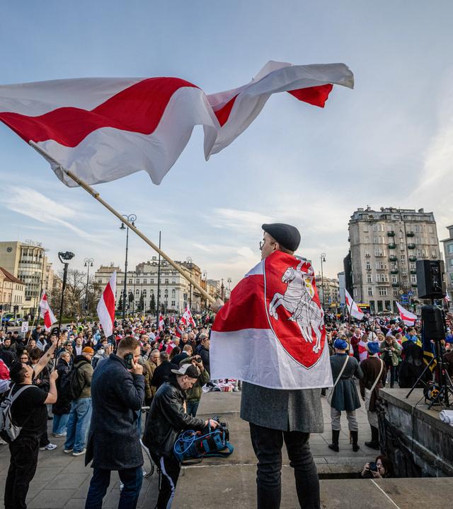 Auf einem großen Platz in einer Großstadt haben sich zahlreiche Menschen mit weiß-rot gestreiften Flaggen versammelt.