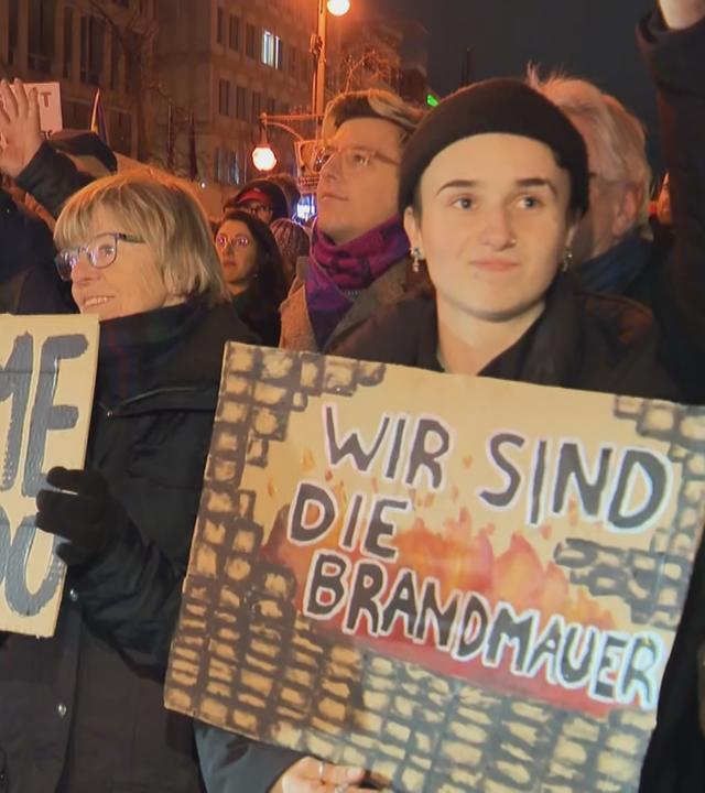 Menschen bei Demo gegen Migrationspolitik in Wiesbaden halten Schilder mit den Aufschriften "Shame on You", "Shame on Merz" und "Wir sind die Brandmauer". 