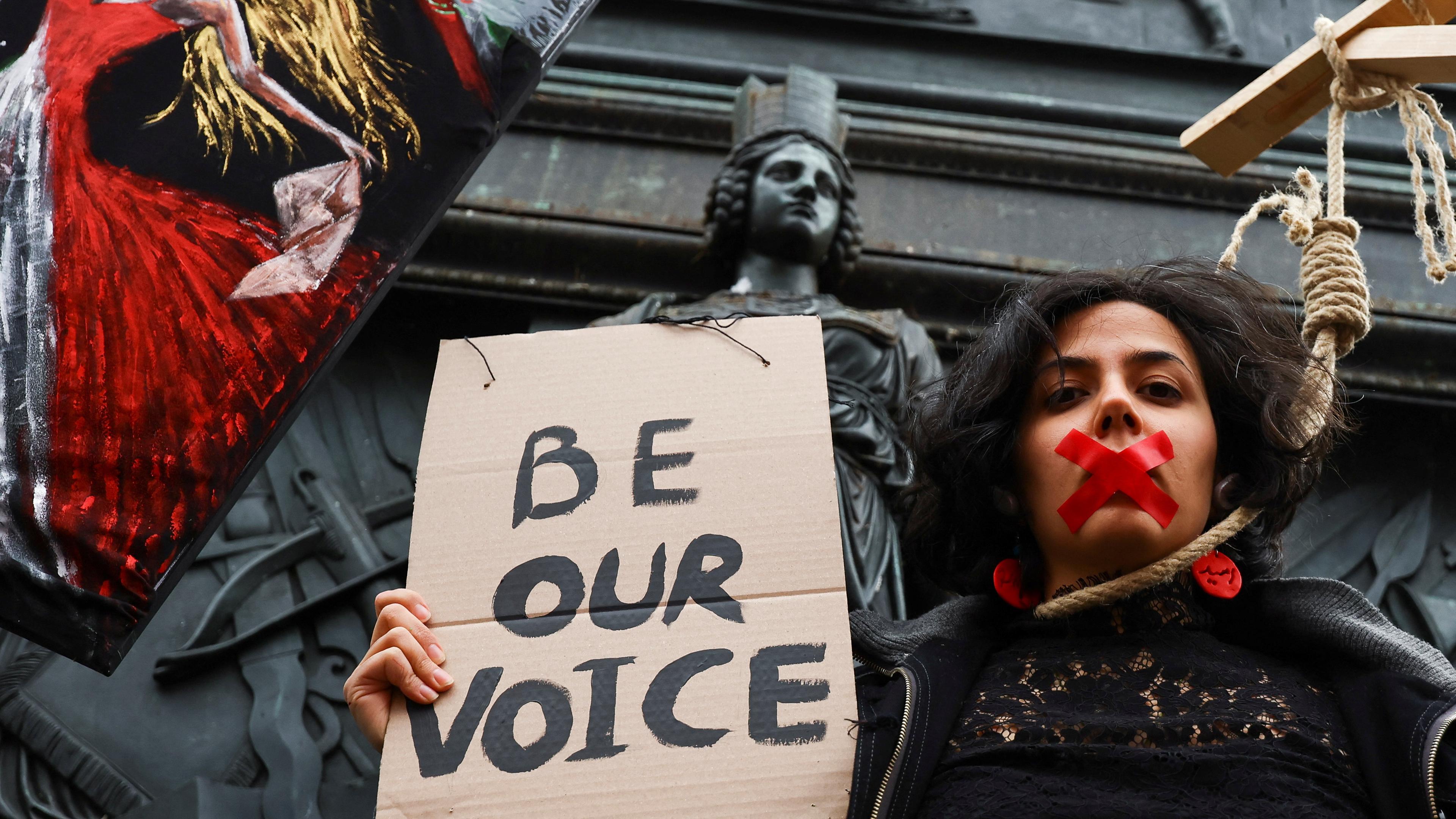 Eine Unterstützerin des iranischen Protests trägt einen Galgenstrick um den Hals und hat den Mund mit rotem Band verklebt. Sie hält ein Schild mit der Aufschrift "Be our voice" - zu Deutsch "Seid unsere Stimme".
