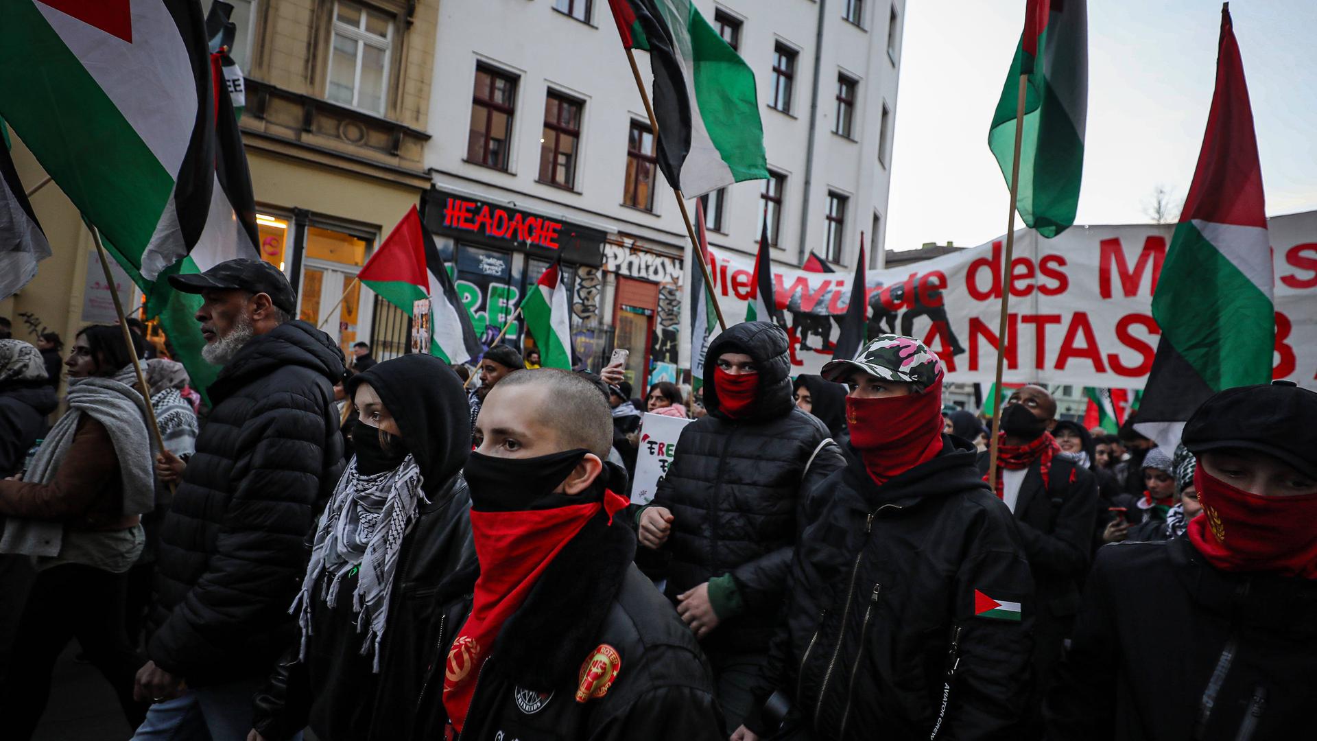 Man sieht eine Demonstration mit maskierten Menschen. Sie halten Flaggen und Banner.