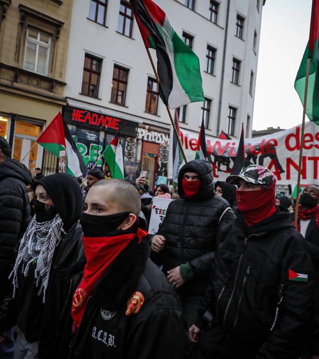 Man sieht eine Demonstration mit maskierten Menschen. Sie halten Flaggen und Banner.