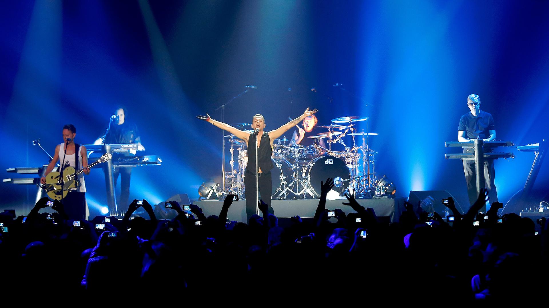 Die Band Depeche Mode in der O2 Arena in Berlin, bei ihrem Auftritt 2013. In der Mitte der blau beleuchteten Bühne Dave Gahan im Spotlight mit ausgebreiteten Armen.