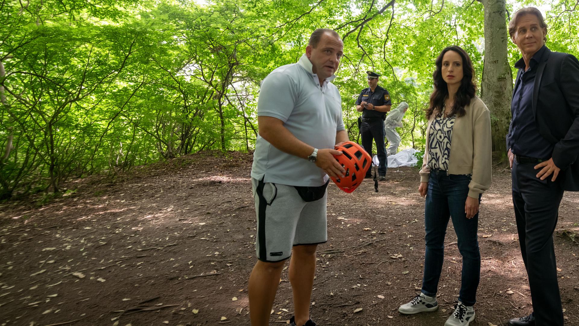 "Der Alte - Abstiegsangst": Simon Rothe (Urs Rechn) steht mit einem Fahrradhelm in der Hand im Wald. Annabell Lorenz (Stephanie Stumph) und Caspar Bergmann (Thomas Heinze) stehen daneben. Alle drei schauen in die Ferne. Im Hintergrund steht ein Polizist bei der Spurensicherung.