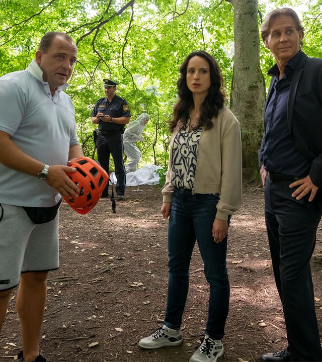 "Der Alte - Abstiegsangst": Simon Rothe (Urs Rechn) steht mit einem Fahrradhelm in der Hand im Wald. Annabell Lorenz (Stephanie Stumph) und Caspar Bergmann (Thomas Heinze) stehen daneben. Alle drei schauen in die Ferne. Im Hintergrund steht ein Polizist bei der Spurensicherung.