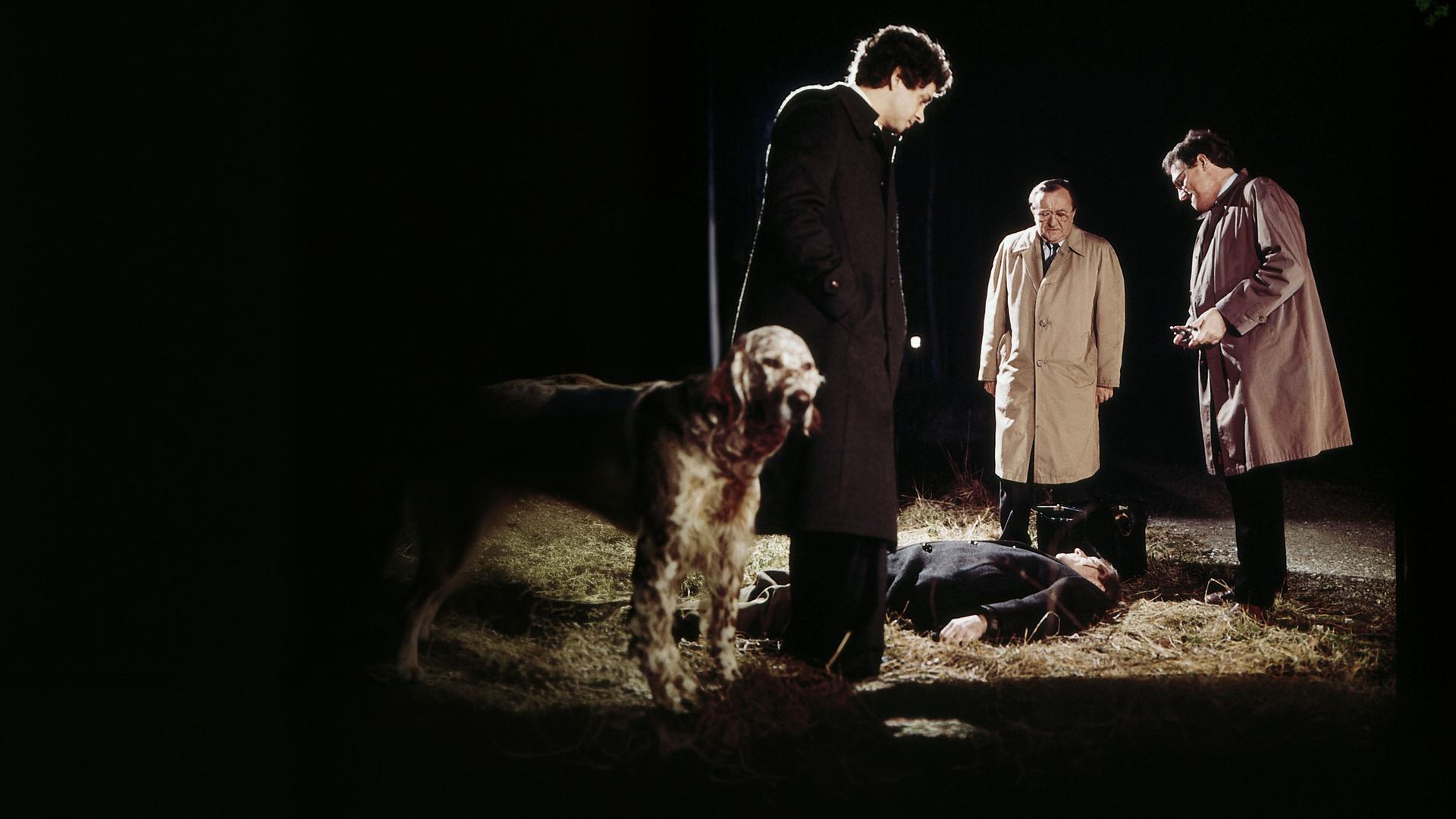 Hauptkommissar Köster (Siegfried Lowitz), Heymann (Michael Ande) und ein dritter Kommissar (Henning Schlüter) stehen vor einem liegenden Mann (Günther Ungeheuer) an einer Aue. Links im Bild steht ein Hund mit hellem Fell. Die drei Kommissare unterhalten sich und schauen nach unten auf den liegenden Toten.