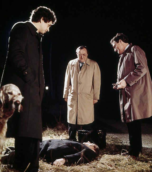Hauptkommissar Köster (Siegfried Lowitz), Heymann (Michael Ande) und ein dritter Kommissar (Henning Schlüter) stehen vor einem liegenden Mann (Günther Ungeheuer) an einer Aue. Links im Bild steht ein Hund mit hellem Fell. Die drei Kommissare unterhalten sich und schauen nach unten auf den liegenden Toten.