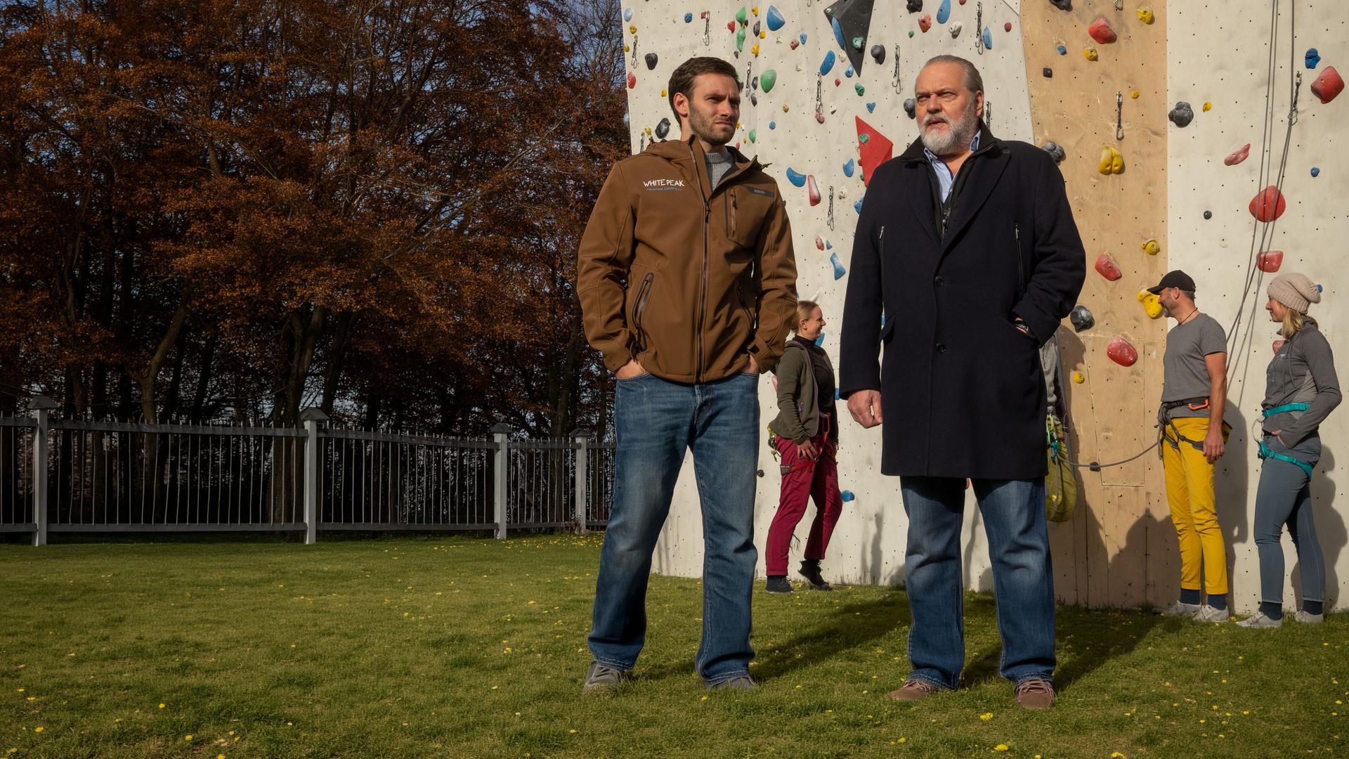 "SOKO Leipzig - Freier Fall": Eric Du Bois (Jonathan Müller) steht mit Hauptkommissar Richard Voss (Jan-Gregor Kremp) im Freien vor einer Kletterwand. Im Hintergrund stehen einige Personen, die sich zum Klettern getroffen haben.