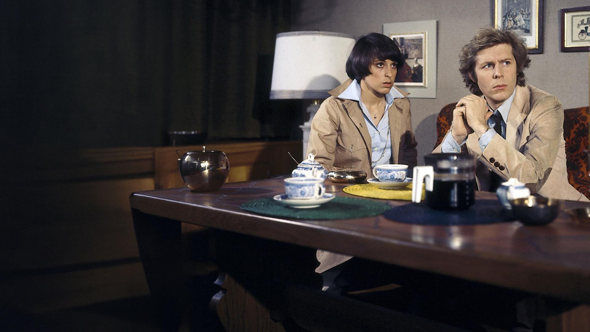 Eva Ströbel (Brigitta Furgler) und Helmut Staufen (Sigmar Solbach) sitzen in einem Wohnzimmer an einem gedeckten Tisch. Eva sitzt oberhalb und Helmut hinter dem Tisch. Sie unterhalten sich. Helmut gibt seine Hände zusammen und schaut nach links.