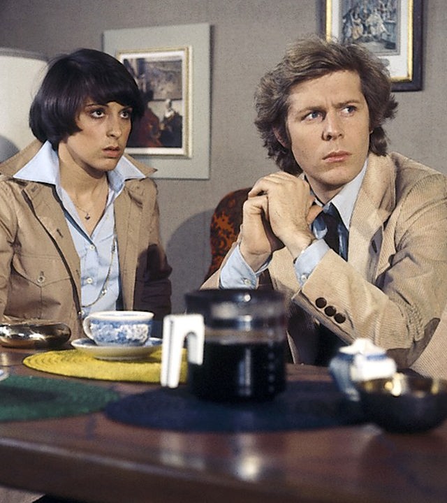 Eva Ströbel (Brigitta Furgler) und Helmut Staufen (Sigmar Solbach) sitzen in einem Wohnzimmer an einem gedeckten Tisch. Eva sitzt oberhalb und Helmut hinter dem Tisch. Sie unterhalten sich. Helmut gibt seine Hände zusammen und schaut nach links.
