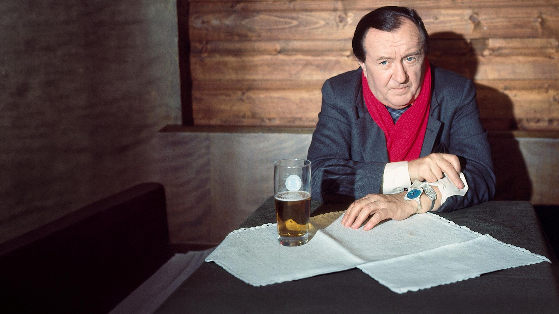 "Der Alte - Der Alte schlägt zweimal zu": Hauptkommissar Köster (Siegfried Lowitz) sitzt an einem mit einem Glas Bier gedeckten Tisch und schaut wartend nach vorne. Er hat auch den Ärmel zurück gekrempelt, an dem eine Uhr zu sehen ist.