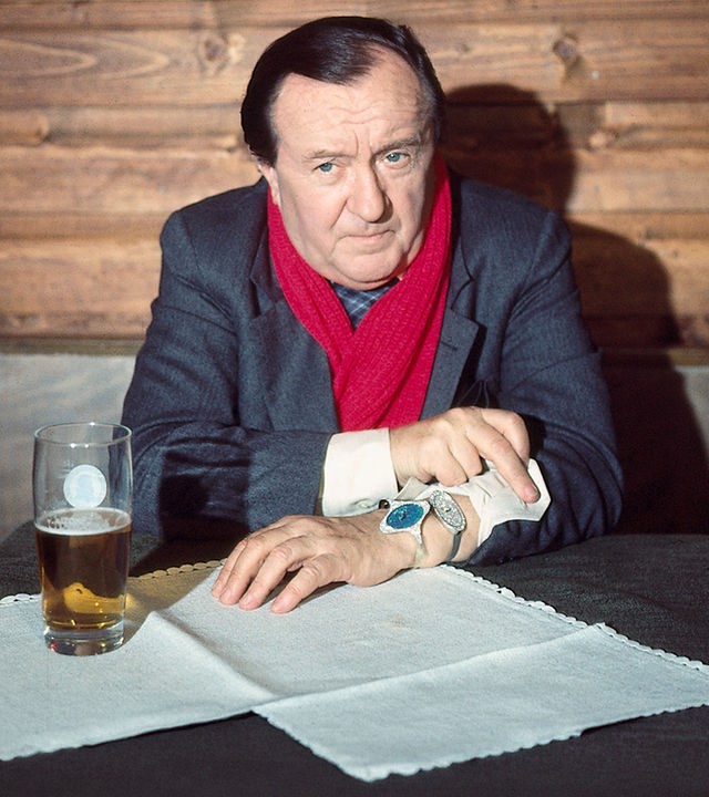 "Der Alte - Der Alte schlägt zweimal zu": Hauptkommissar Köster (Siegfried Lowitz) sitzt an einem mit einem Glas Bier gedeckten Tisch und schaut wartend nach vorne. Er hat auch den Ärmel zurück gekrempelt, an dem eine Uhr zu sehen ist.