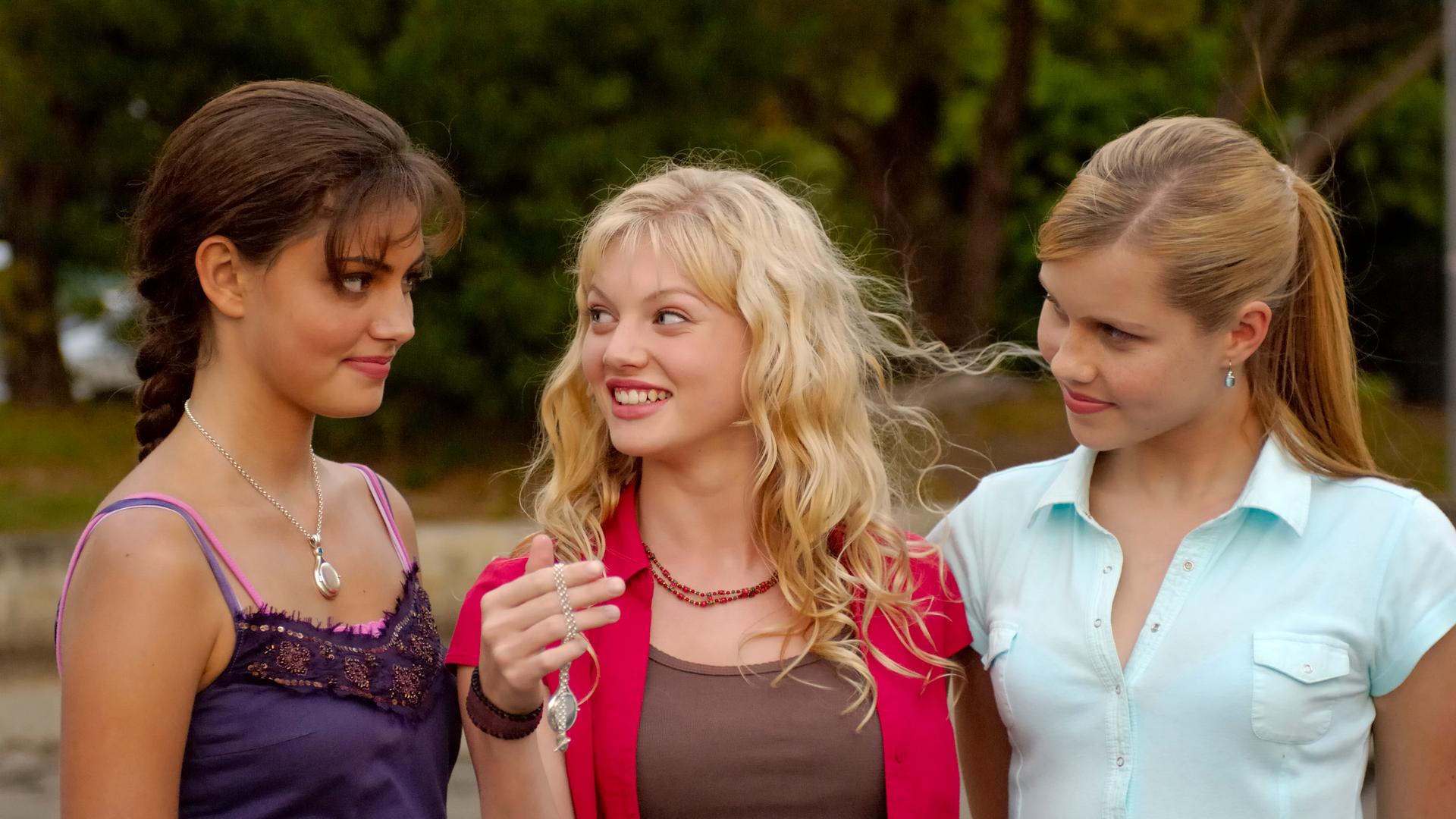 Rikki hält den Anhänger in ihrer Hand und lächelt. Cleo und Emma lächeln ebenfalls.