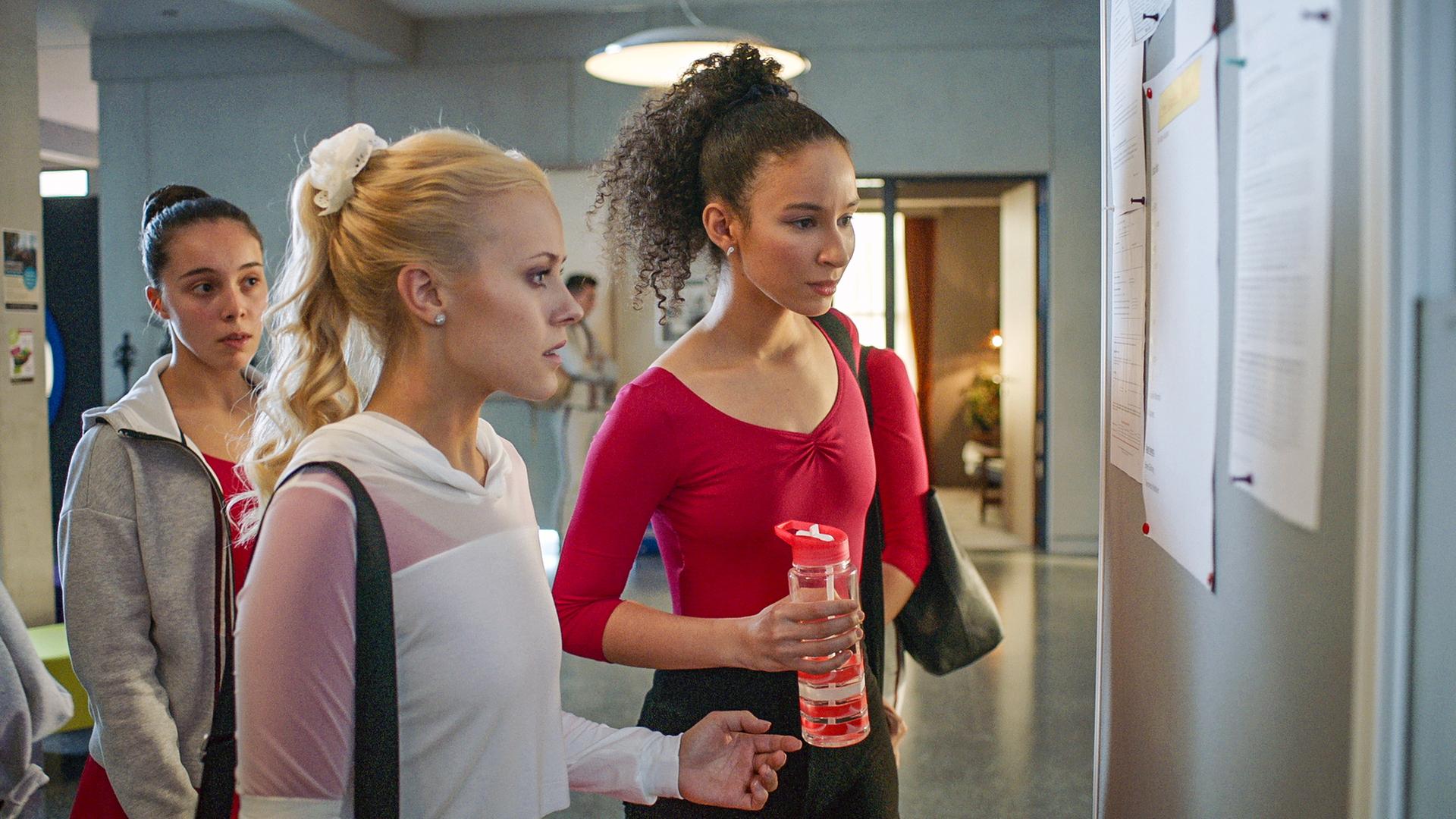 Lena (Mitte) und Ines (rechts) sehen sich im Flur den Aushang mit den Rollen für die "Carte Blanche"-Show an. Sie wirken geschockt.