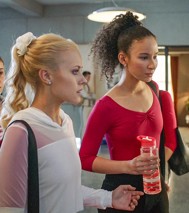 Lena (Mitte) und Ines (rechts) sehen sich im Flur den Aushang mit den Rollen für die "Carte Blanche"-Show an. Sie wirken geschockt.