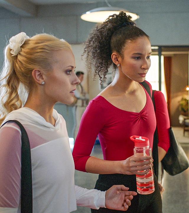 Lena und Ines sehen sich im Flur den Aushang mit den Rollen für die "Carte Blanche"-Show an. Sie wirken geschockt.