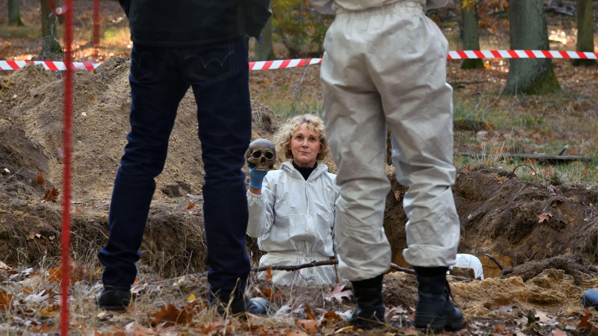 "Die Spezialisten - Der Bankier": Maria Kaschke (Rosa Enskat) steht in einem großen Erdloch in einem Wald. Sie trägt einen weißen Overall der Spurensicherung. In ihrer rechten Hand hält sie einen Schädel. Sie schaut zu Dr. Julia Löwe (Alina Levshin) und Hauptkommissar Henrik Mertens (Matthias Weidenhöfer) und grinst. Beide stehen hinter der Absperrung. Man sieht die beiden von hinten nur bis zur Mitte des Rückens, Schulterbereich und Kopf sind außerhalb des Fotos.