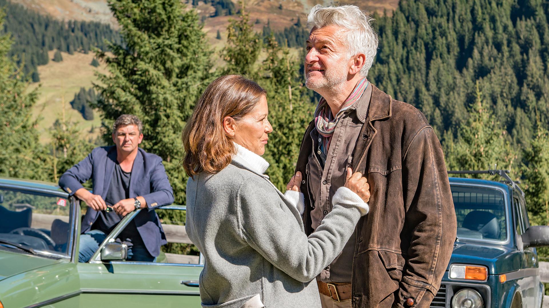 Martin (Hans Sigl) steht an seinem Auto und beobachtet seinen Patient Arno Moosbach (Dominic Raacke) und Katrin (Angela Roy), die eng und lächelnd beieinander stehen.