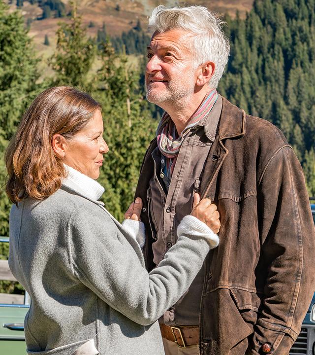 Martin (Hans Sigl) steht an seinem Auto und beobachtet seinen Patient Arno Moosbach (Dominic Raacke) und Katrin (Angela Roy), die eng und lächelnd beieinander stehen.