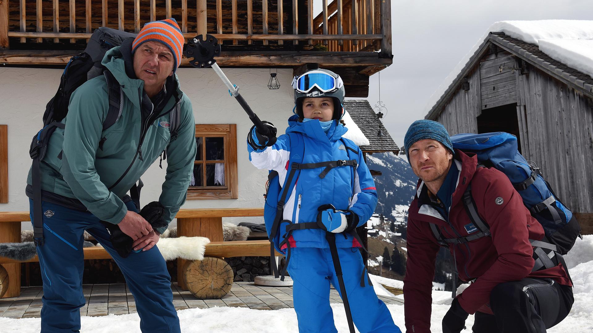Vor einer Hütte steht Flora (Yuna Bennet) im Schnee. Sie trägt Skibekleidung und zeigt mit einem ihrer Skistöcke in die Ferne. Martin (Hans Sigl) steht links, sein Bruder Hans (Heiko Ruprecht) kniet rechts neben ihr. Beide blicken fragend in die Ferne.