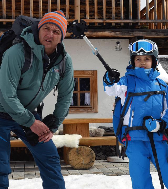 Vor einer Hütte steht Flora (Yuna Bennet) im Schnee. Sie trägt Skibekleidung und zeigt mit einem ihrer Skistöcke in die Ferne. Martin (Hans Sigl) steht links, sein Bruder Hans (Heiko Ruprecht) kniet rechts neben ihr. Beide blicken fragend in die Ferne.