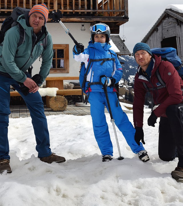 Vor einer Hütte steht Flora (Yuna Bennet) im Schnee. Sie trägt Skibekleidung und zeigt mit einem ihrer Skistöcke in die Ferne. Martin (Hans Sigl) steht links, sein Bruder Hans (Heiko Ruprecht) kniet rechts neben ihr. Beide blicken fragend in die Ferne.