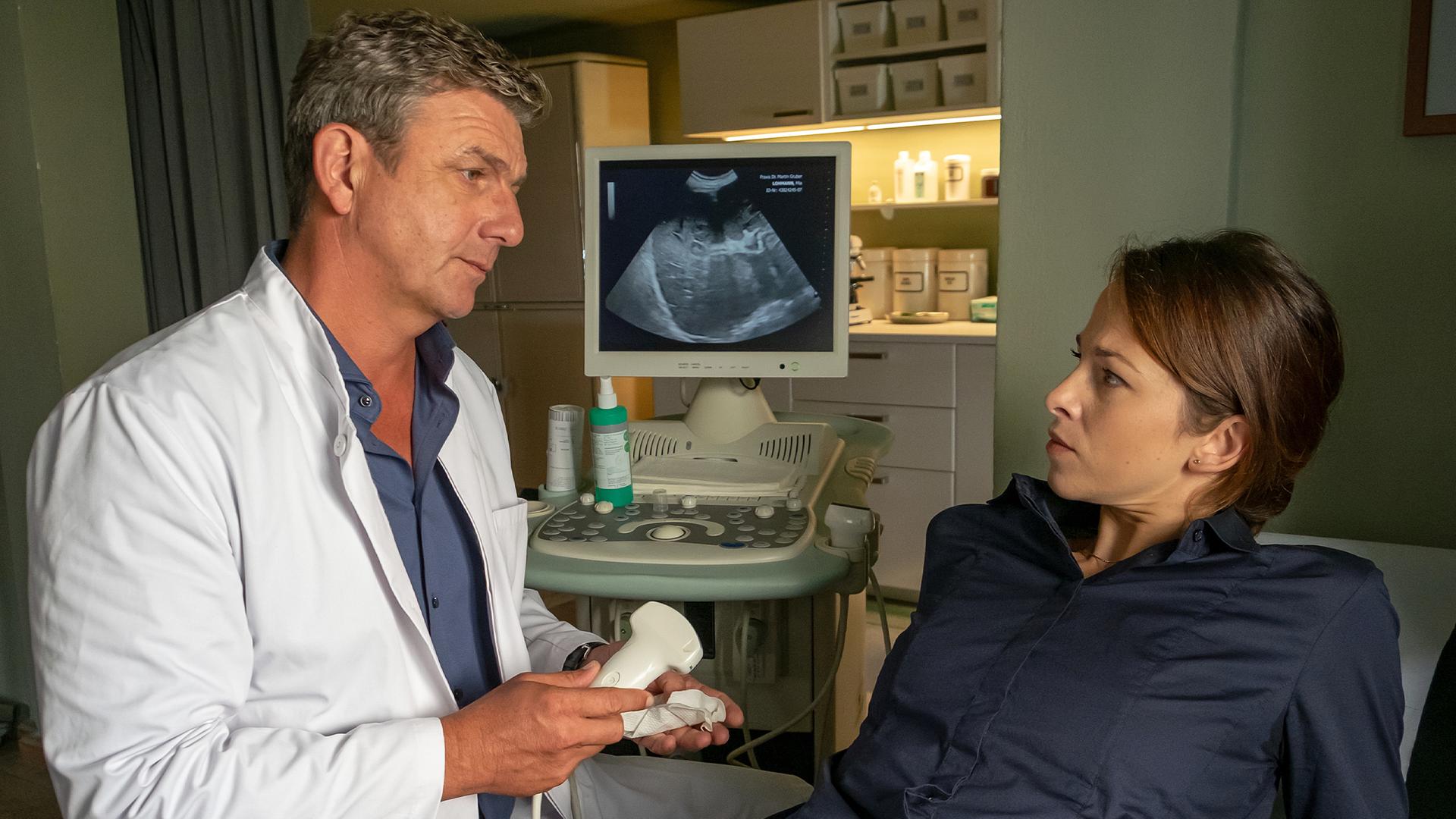 In der Arztpraxis: Dr. Martin Gruber (Hans Sigl) sitzt, ein Ultraschallgerät haltend, neben Mia Lohmann (Marie Hacke), die auf einer Untersuchungsliege vor ihm liegt. Beide sehen sich ernst an.