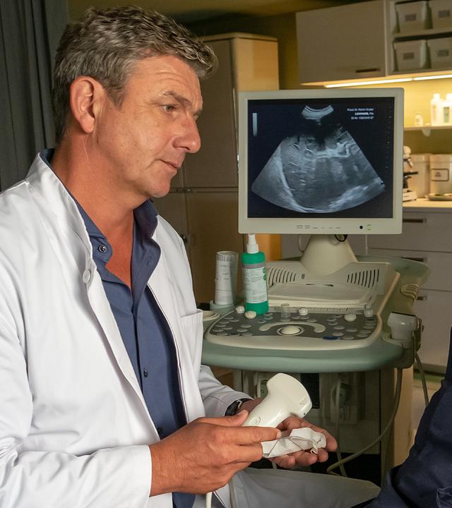 In der Arztpraxis: Dr. Martin Gruber (Hans Sigl) sitzt, ein Ultraschallgerät haltend, neben Mia Lohmann (Marie Hacke), die auf einer Untersuchungsliege vor ihm liegt. Beide sehen sich ernst an.