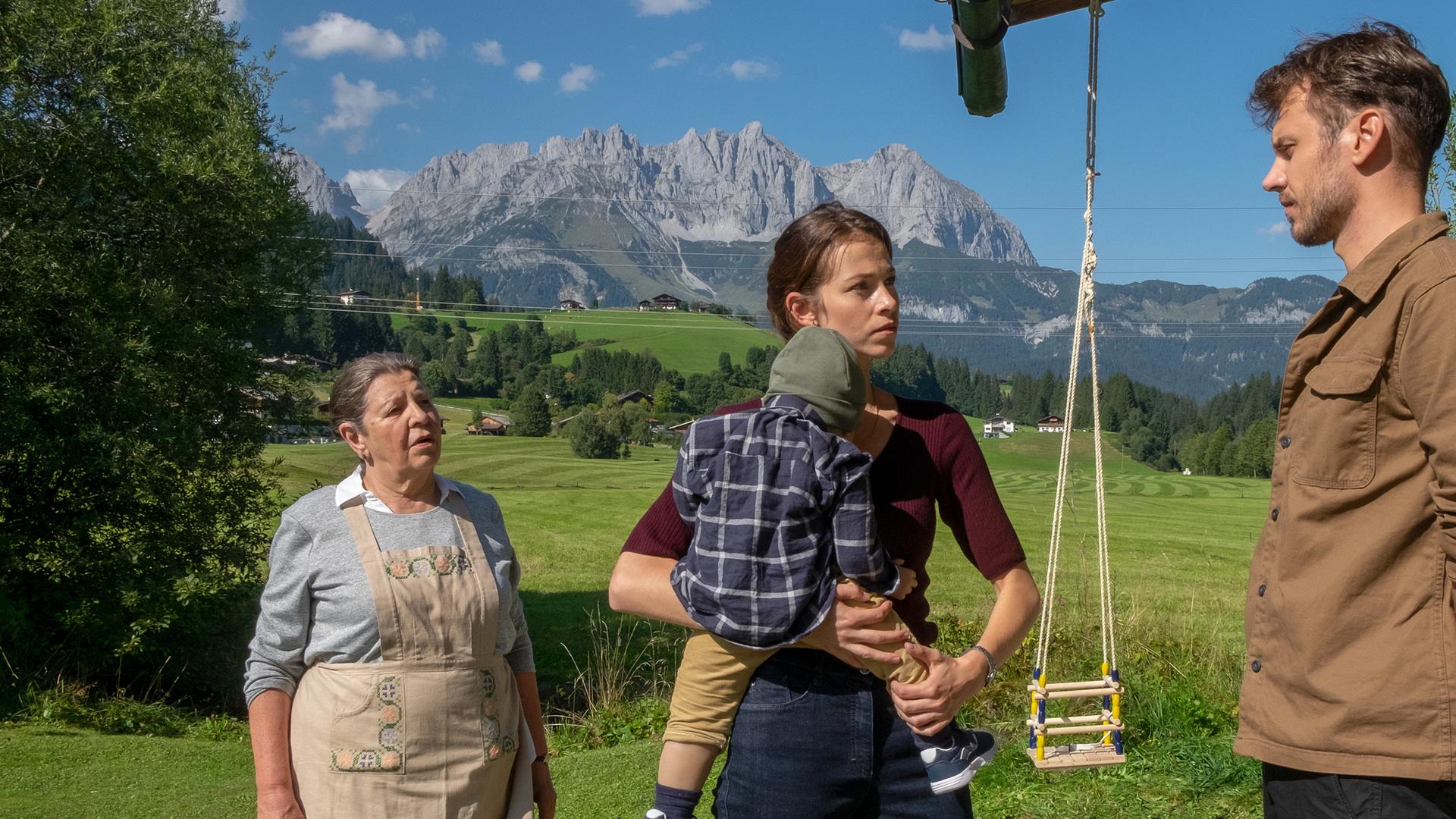 Mia Lohmann (Marie Hacke) trägt Baby Felix (Emilian Koller) auf ihrem Arm und steht zwischen Elsa (Monika Manz) und David Berger (Michael Glantschnig) vor ihrem Haus. Im Hintergrund eine sonnige Bergkulisse.