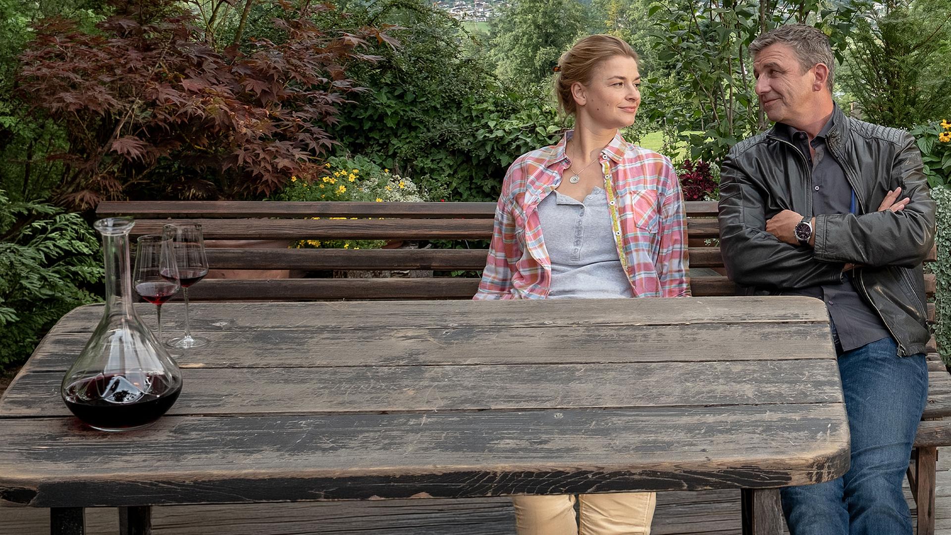Vor einer idyllischen Landschaft und umringt von Blumenkübeln sitzen Anne (Ines Lutz) und Marin (Hans Sigl) auf einer Holzbank nebeneinander. Sie lächeln sich an.