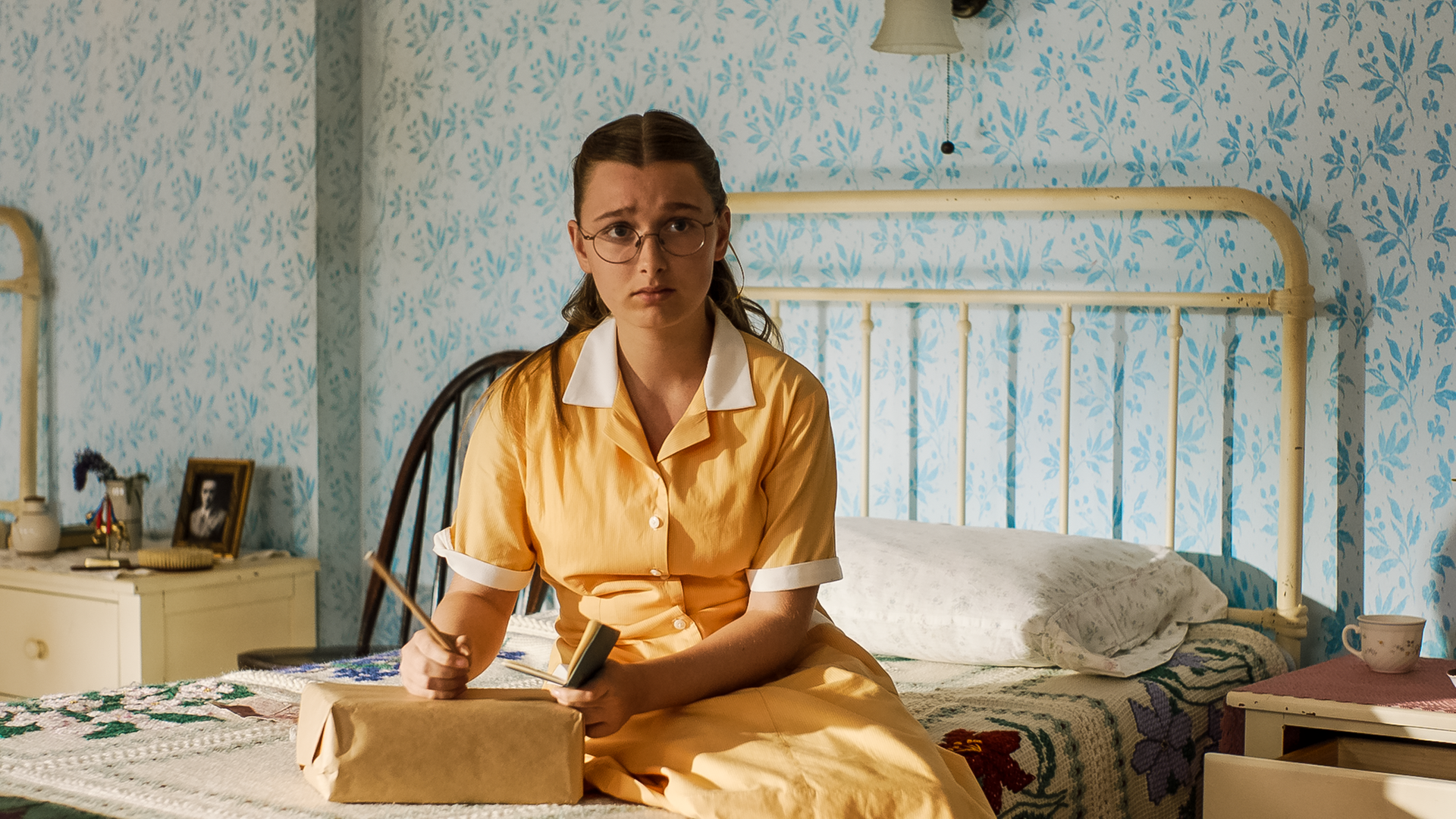 Mary-Lou sitzt auf ihrem Bett im Schlafsaal. Vor ihr liegt ein braunes Paket, auf das sie eine Adresse etwas schreibt. Sie wirkt besorgt.