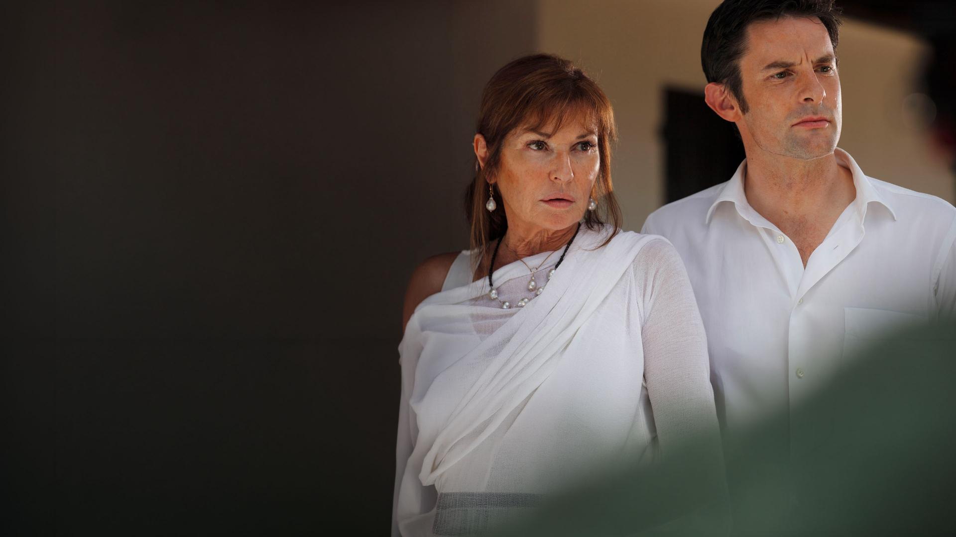 "Death in Paradise - Der blutige Finger": Nahaufnahme Nicole Seymour (Stephanie Beacham) und Alex Seymour (Tom Ward). Beide tragen weiße Oberteile und scheinen jemanden anzusehen.