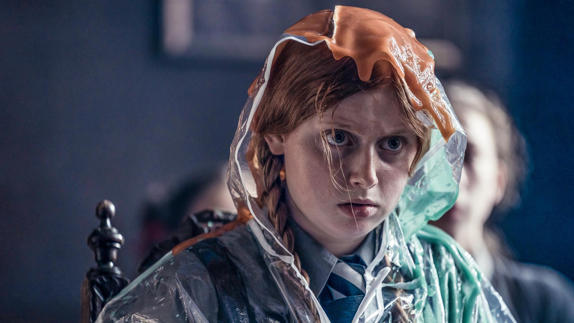 Mildred in einer misslichen Lage: Sie sitzt mit mehreren Karteikarten in ihren Händen auf einem Stuhl und schaut dabei unglücklich nach vorne. Sie hat zum Glück einen transparenten Plastikumhang mit Kapuze an, denn über ihren Kopf laufen zwei hässliche, schleimige Flüssigkeiten in orange und grün.