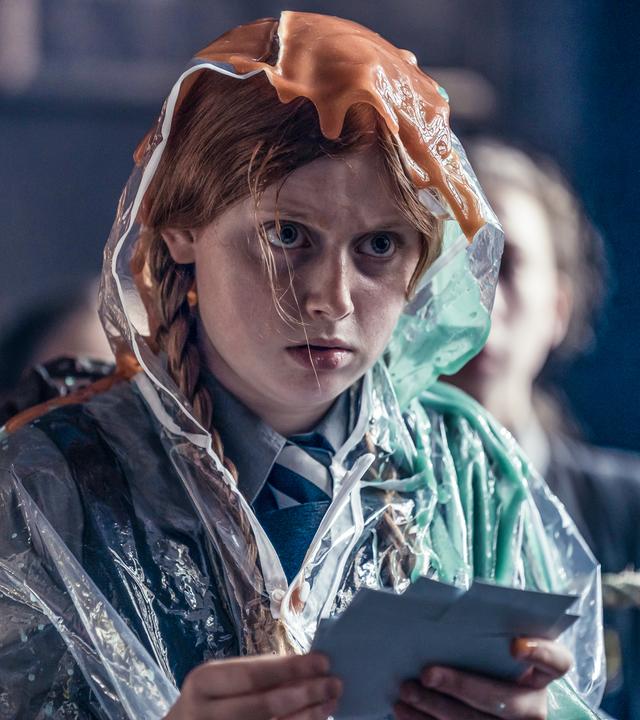 Mildred in einer misslichen Lage: Sie sitzt mit mehreren Karteikarten in ihren Händen auf einem Stuhl und schaut dabei unglücklich nach vorne. Sie hat zum Glück einen transparenten Plastikumhang mit Kapuze an, denn über ihren Kopf laufen zwei hässliche, schleimige Flüssigkeiten in orange und grün.