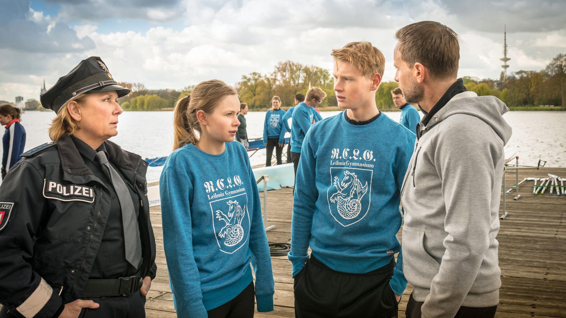 "Notruf Hafenkante - Der doppelte Viktor": Auf einem großen Holzsteg stehen die Polizistin Claudia Fischer (Janette Rauch), Sinje Kästner (Emilia Bernsdorf), Fabian Frenz (Lukas Zumbrock) und Uwe Kästner (Julian Weigend) in einer Gruppe beisammen und unterhalten sich.