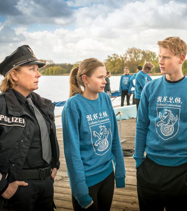 "Notruf Hafenkante - Der doppelte Viktor": Auf einem großen Holzsteg stehen die Polizistin Claudia Fischer (Janette Rauch), Sinje Kästner (Emilia Bernsdorf), Fabian Frenz (Lukas Zumbrock) und Uwe Kästner (Julian Weigend) in einer Gruppe beisammen und unterhalten sich.
