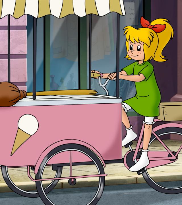 Bibi fährt mit einem rosafarbenen Eisfahrrad auf der Straße an Schaufenstern vorbei. Ihr Besen liegt auf dem Speiseeisbehälter vor ihr.