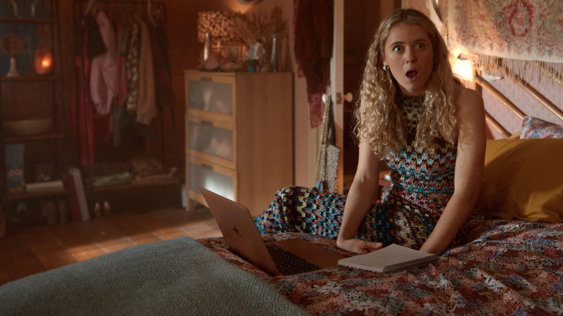 "Extraordinary - Der Exorzismus der Carrie Jackson": Carrie (Sofia Oxenham) sitzt auf einem Bett in einem gemütlich eingerichteten Schlafzimmer, vor ihr liegen ein Laptop und ein Notizbuch. Mit offenem Mund blickt sie erstaunt zur Seite.