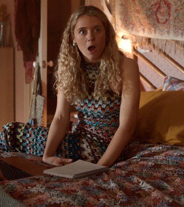 "Extraordinary - Der Exorzismus der Carrie Jackson": Carrie (Sofia Oxenham) sitzt auf einem Bett in einem gemütlich eingerichteten Schlafzimmer, vor ihr liegen ein Laptop und ein Notizbuch. Mit offenem Mund blickt sie erstaunt zur Seite.