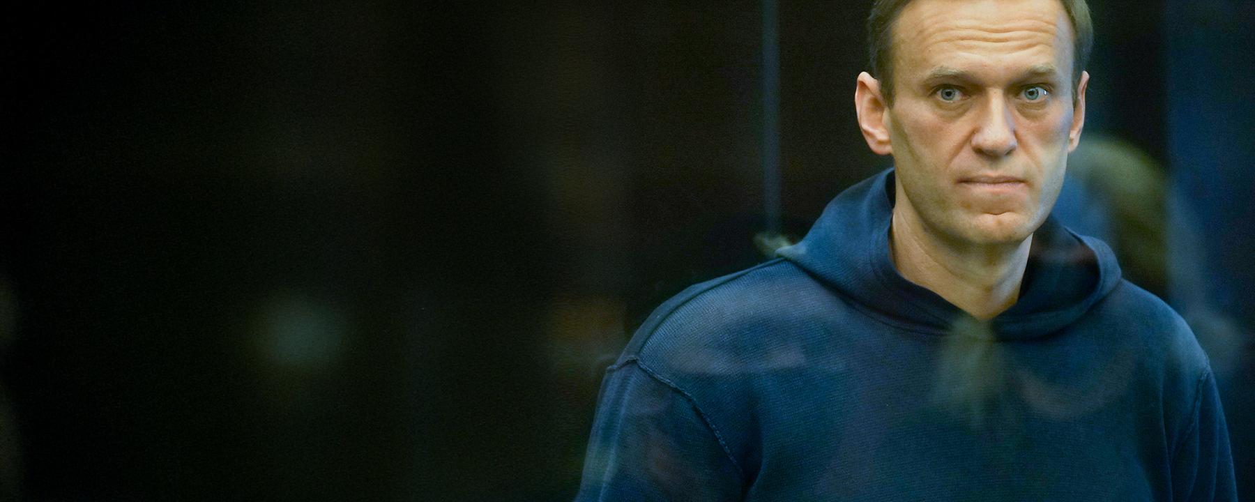 Alexey Nawalny steht im Käfig im Gericht, bekleidet mit einem dunkelblauen Hoody und schaut direkt durch die Glasscheibe in die Kamera.