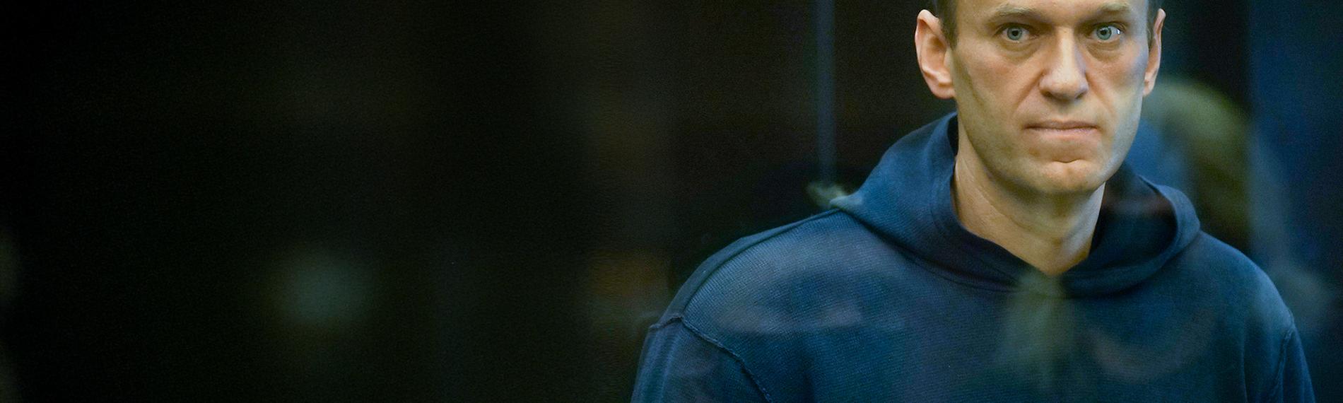 Alexey Nawalny steht im Käfig im Gericht, bekleidet mit einem dunkelblauen Hoody und schaut direkt durch die Glasscheibe in die Kamera.