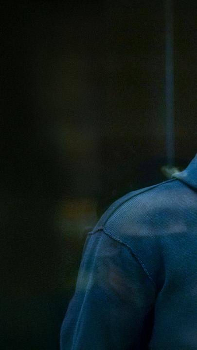 Alexey Nawalny steht im Käfig im Gericht, bekleidet mit einem dunkelblauen Hoody und schaut direkt durch die Glasscheibe in die Kamera.